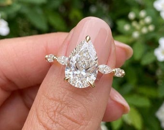 May include: A gold ring featuring a large pear-shaped center stone held by prongs, flanked by two smaller marquise-cut stones on each side of the band. The ring has a polished gold finish and a delicate, slender band design with clear, faceted stones.