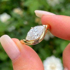 May include: A gold ring featuring a single marquise-cut clear gemstone set in a smooth, polished yellow gold band that curves around the base of the stone. The design highlights the elongated shape of the faceted center stone and the warm tone of the metal.