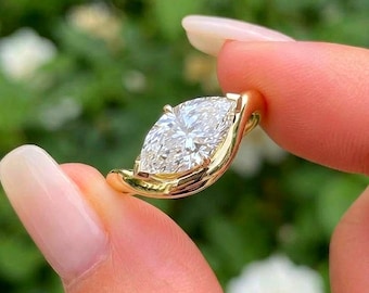 May include: A gold ring featuring a single marquise-cut clear gemstone set in a smooth, polished yellow gold band that curves around the base of the stone. The design highlights the elongated shape of the faceted center stone and the warm tone of the metal.