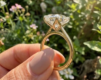 May include: A gold ring with a large, round, clear gemstone. The ring is held up against a background of green foliage and small white and pink flowers. The ring's band is a simple, polished gold.