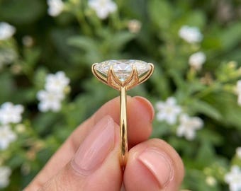 May include: A gold ring featuring a large, clear, oval-cut gemstone held in a six-prong setting. The band is thin, smooth, and polished gold, showcasing the side profile of the stone and the prong structure against a blurred green background.