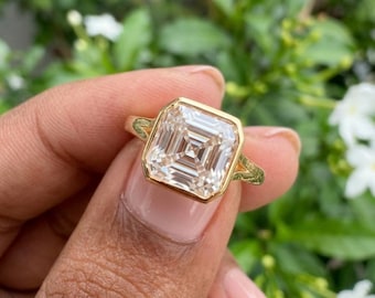 May include: A gold ring featuring a large, square-cut, clear gemstone with a step-cut facet pattern. The stone is set in a polished gold bezel setting that transitions into a simple, smooth gold band, showcasing a bright, reflective appearance.
