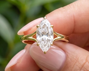 May include: A marquise-cut clear gemstone ring set in a polished yellow gold band. The ring features a split-shank design where the band divides as it reaches the central stone, which is held in place by a prong setting with a pointed tip.