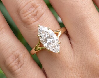 May include: A gold ring featuring a large, marquise-cut clear gemstone held in a prong setting. The band has a split shank design that tapers toward the center stone, creating a sleek and polished appearance on the finger.