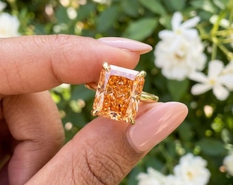 May include: A ring featuring a large, rectangular, radiant-cut orange gemstone set in a yellow gold band with four prongs. The stone displays a vibrant, saturated orange hue with multiple facets that reflect light, creating a brilliant, sparkling appearance.