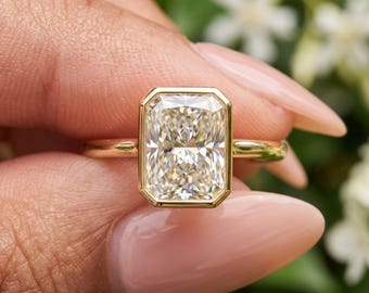May include: A radiant-cut clear gemstone set in a yellow gold bezel setting on a thin, polished yellow gold band. The rectangular stone features multiple facets that reflect light, creating a brilliant appearance within the smooth metal frame.