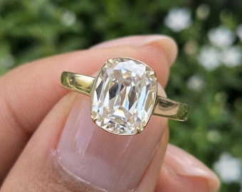 May include: A gold ring featuring a large, cushion-cut, clear gemstone set in a four-prong setting. The band is smooth and polished, showcasing a classic design with a prominent, faceted center stone that reflects light across its surface.
