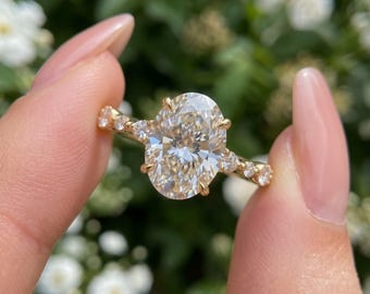 May include: A gold ring featuring a large oval-cut center stone held by four prongs, flanked by three smaller round-cut stones on each side of the band. The metal band has a polished finish, and the stones are clear and faceted to reflect light.