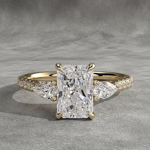 Può includere: Anello di fidanzamento in oro con diamante taglio rettangolare. L'anello presenta una grande pietra centrale rettangolare, affiancata da due diamanti a forma di pera. La fascia è impreziosita da pietre più piccole.