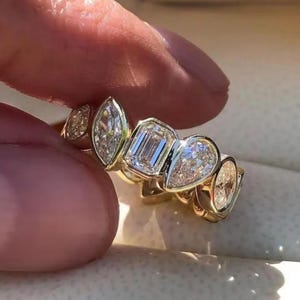 Può includere: Un anello d'oro con una serie di diamanti di forme diverse. I diamanti includono tagli a marquise, smeraldo, pera e ovali. L'anello è tenuto dalle dita di una persona, mostrando l'intricato design e la brillantezza delle pietre preziose.