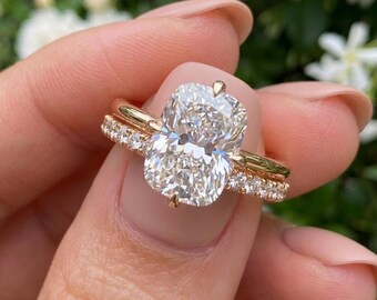 May include: A gold engagement ring with a large, oval-cut diamond as the centerpiece. The band is also gold and features smaller diamonds. The ring is held up against a blurred green and white floral background.