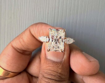 May include: A gold ring with a large, rectangular, clear gemstone as the centerpiece, flanked by two smaller pear-shaped stones. The ring is held up by a person's fingers, with another gold band visible on a finger.