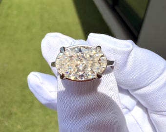 May include: A silver-toned ring featuring a large, horizontally oriented oval-cut clear gemstone held in place by four prominent prongs. The stone exhibits multiple facets that reflect light, creating a bright, sparkling appearance against the metal band.