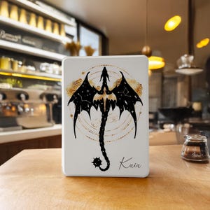 Puede incluir: Funda rectangular blanca con un diseño de dragón negro con detalles de constelaciones. El dragón tiene alas extendidas y una larga cola. También se ven detalles dorados y el nombre "Kaia".