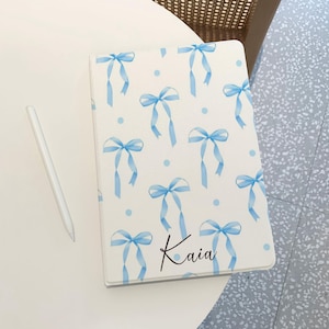 Puede incluir: Una cubierta de cuaderno blanca con un patrón de lazos azules y el nombre "Kaia" escrito en cursiva.