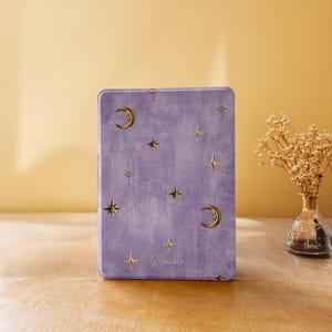 May include: A rectangular purple book cover with a textured finish featuring gold foil illustrations of crescent moons and various star shapes scattered across the surface. The word "Karin" is written in a delicate cursive script near the bottom.