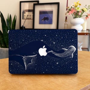 Puede incluir: Una funda para portátil azul marino con el logotipo blanco de Apple y un diseño de cielo estrellado. El diseño incluye una manta raya y un tiburón ballena, con estrellas blancas.