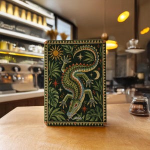 Puede incluir: Un cuaderno rectangular con cubierta verde oscuro que presenta una ilustración central de un cocodrilo rodeado de hojas tropicales, un sol, una luna creciente y estrellas. La palabra Gaia está escrita en cursiva blanca abajo.