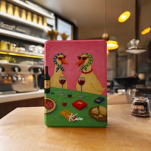 Könnte beinhalten: Eine rechteckige Metallbox mit rosa Hintergrund, die zwei Enten mit Sonnenbrillen und Schals beim Weintrinken zeigt. Ein grüner Tisch zeigt Weingläser, eine Flasche, Snacks und den Namen "Kaia".