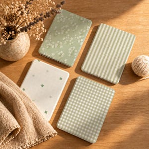 May include: Four rectangular notebooks with rounded corners. One has a green floral pattern, one has green vertical stripes, one has a white grid with small green clover icons, and one has a green and white gingham check pattern.