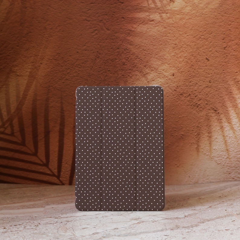 May include: A brown tablet case with a white polka dot pattern. The case is rectangular and has a folding cover. The background is a textured brown wall with a shadow of a leaf.