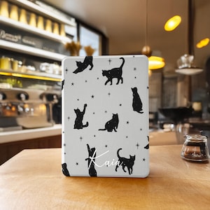 Pode incluir: Uma capa retangular cinza claro com silhuetas de gatos pretos e estrelas. Os gatos estão em várias poses. O nome "Kaia" está escrito em itálico. A capa está em uma superfície de madeira.
