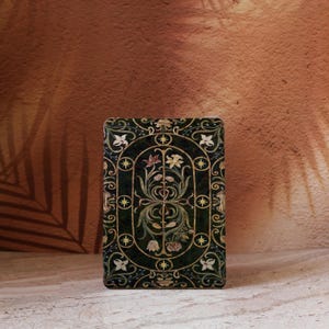 Puede incluir: Una caja rectangular verde oscuro con un diseño floral ornamentado. El diseño presenta detalles dorados, formas de estrella y flores estilizadas dentro de un marco ovalado. La caja tiene esquinas redondeadas y un aspecto texturizado.