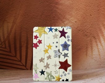 Può includere: Una custodia per tablet color crema adornata con una varietà di disegni a stella colorati. Le stelle sono in tonalità oro, rosso, blu e verde, con alcuni motivi intricati. La custodia è rettangolare ed è progettata per proteggere un tablet.