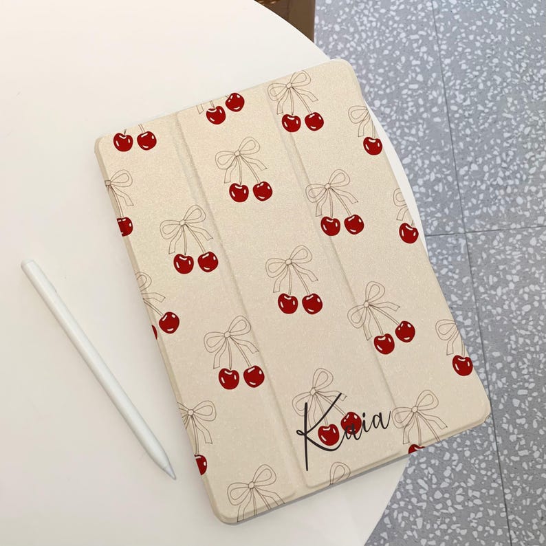 May include: A beige iPad case with a repeating pattern of red cherries and black bows. The case has the name "Kalia" written in black cursive on the bottom right corner.