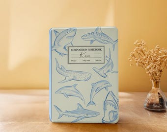 Puede incluir: Un cuaderno de composición con una cubierta azul claro con ilustraciones azules de varias especies de tiburones y delfines. El cuaderno tiene el texto "COMPOSITION NOTEBOOK Kaia" y mide 19 x 14 cm.