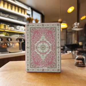 Könnte beinhalten: Ein rechteckiger Gegenstand mit einem rosa-beigen Muster, das an einen Teppich erinnert. Das Design weist florale Motive auf. Der Gegenstand befindet sich auf einer Holzoberfläche. Das Wort "Kaviri" ist unten rechts in Kursivschrift geschrieben.