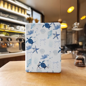 Könnte beinhalten: Eine weiße Tablethülle mit einem blauen und weißen Meeresmuster. Das Muster zeigt Meeresschildkröten, Seesterne, Muscheln und florale Akzente. Die Hülle trägt den Namen "Kira" aufgedruckt.