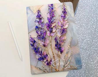 Puede incluir: Un cuaderno decorativo con un diseño floral que presenta flores moradas y lavanda con detalles dorados. El cuaderno tiene un fondo nacarado y la palabra "Kaia" está escrita en la portada. Un lápiz óptico blanco está a la izquierda.