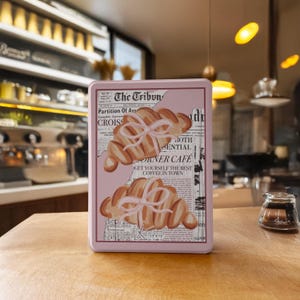 May include: A pink rectangular tin featuring two croissants tied with ribbons. The design includes newspaper-style text and the words "The Tribun" and "ORNER CAFE". The tin is on a wooden surface, suggesting a cafe setting.