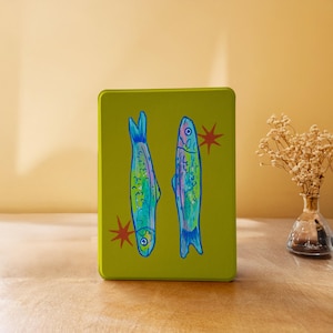Puede incluir: Una lata rectangular verde oliva con una ilustración dibujada a mano de dos peces en tonos azules y verdes. Estallidos de estrellas rojas flanquean los peces. El fondo es de un color cálido y neutro. La lata es probablemente de metal.