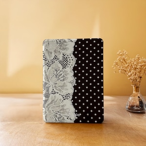 May include: A rectangular e-reader case featuring a split design. The left side displays a white floral lace pattern on a black background, while the right side features a black background with a white polka dot pattern. The name Kaja is printed in white.