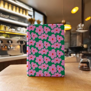 Può includere: Una custodia rettangolare con sfondo verde e un motivo di fiori rosa con centri rossi. La custodia presenta la firma manoscritta "Kaia" in bianco. La custodia è appoggiata su una superficie di legno.