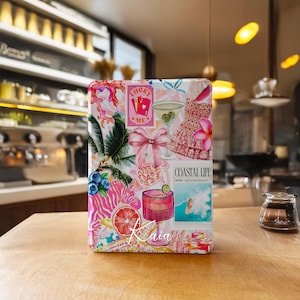 Könnte beinhalten: Eine farbenfrohe, gemusterte Hülle mit einer Collage aus tropischen und Küstenmotiven. Das Design umfasst ein Martiniglas, eine rosa Schleife, ein Kleid und die Worte "Coastal Life". Die Hülle befindet sich auf einer Holzoberfläche.