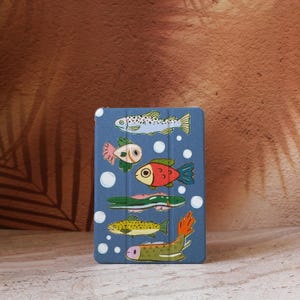 Puede incluir: Una funda azul para tableta con ilustraciones de peces de dibujos animados coloridos. Los peces tienen varias formas y colores, incluyendo rojo, verde, amarillo y blanco, con detalles de burbujas. La funda está diseñada para proteger una tableta.