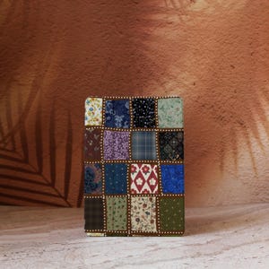 May include: A square, patchwork-style object with a variety of fabric patterns in a grid. The fabrics include floral, plaid, and patterned designs in shades of blue, green, purple, and brown. The edges are trimmed with a brown border and white dots.