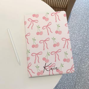 May include: A light pink notebook with a pattern of pink cherries and bows. The name "Kaia" is written in cursive on the bottom right corner. A white stylus pen rests on the table next to the notebook.