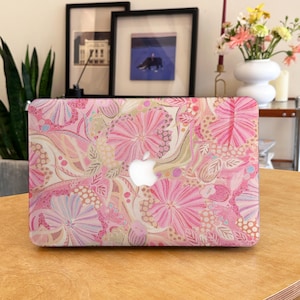 Könnte beinhalten: Eine Laptop-Hülle mit einem lebendigen Blumenmuster in Rosa-, Pfirsich- und Cremetönen. Das Design zeigt stilisierte Blüten, Blätter und gepunktete Texturen auf der gesamten Oberfläche, mit einem zentralen Ausschnitt für das Apple-Logo.