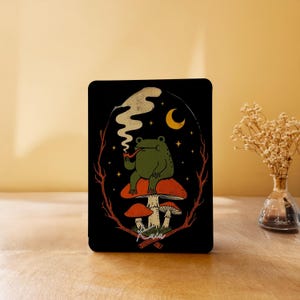 May include: Black rectangular art print featuring a green frog smoking a pipe while seated on a red mushroom. The illustration includes a crescent moon, stars, and the text "Kaia". The background is black with a white smoke trail.