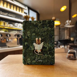 May include: A rectangular e-reader cover featuring a dark green forest foliage pattern with a central illustration of a small brown mouse sitting in a chair reading a newspaper. The name "Kala" is written in white cursive script at the bottom.