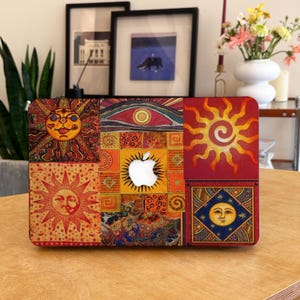 May include: A vibrant laptop case with a patchwork design. It features sun and moon illustrations in orange, red, and blue. The case has a cutout for the Apple logo and is placed on a wooden table.