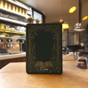 May include: A dark green rectangular e-reader cover featuring a gold-colored line art illustration of a forest with trees, a crescent moon, and an open book at the bottom. The name "Kaia" is written in white cursive script below the open book.