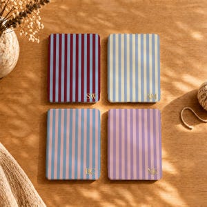 May include: Four rectangular cases featuring vertical stripes in maroon and blue, light blue and cream, light blue and pink, and lavender and pink. Each case displays two gold-colored capital letters in the bottom right corner: SW, AM, BC, and NK.