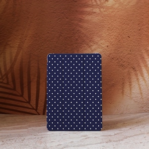 Puede incluir: Una funda para tableta azul marino con un estampado de lunares blancos. La funda es rectangular y tiene una superficie lisa. El fondo es una pared marrón texturizada.