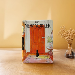 May include: A rectangular tin featuring a vintage "The New Yorker" cover illustration. The artwork depicts a black cat sitting in front of an orange door, with a clock and a table with flowers. The tin has the text "Feb. 4, 1974" and "Price 50 cents".