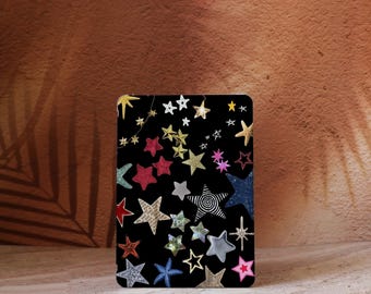 Può includere: Un oggetto rettangolare nero ricoperto da vari disegni di stelle colorate. Le stelle sono di diverse dimensioni, colori e trame, tra cui glitter, fantasia e tinta unita. Lo sfondo è una superficie marrone strutturata.